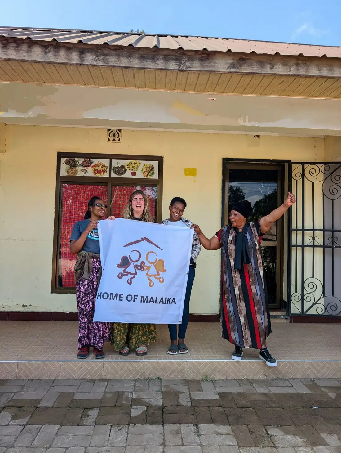 Freiwillige mit Home-of-Malaika-Banner vor dem Restaurant